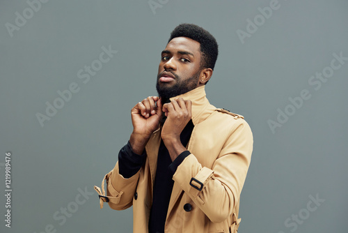 Stylish young Black man in a beige trench coat, confidently adjusting his collar against a gray background, exuding modern elegance and self assurance photo