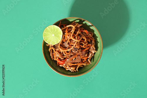 Bowl of tasty Chinese noodles, vegetables, meat and lime on color background
