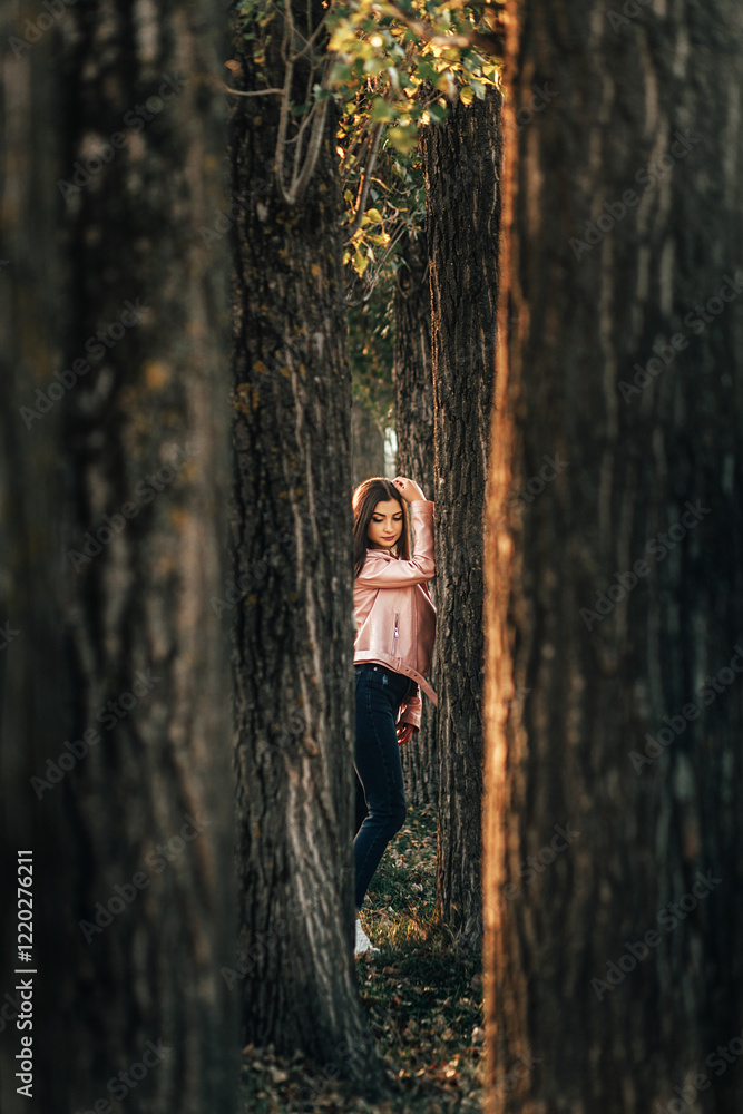 girl standing between rows of trees. She is wearing a pink jacket and dark jeans. The girl tilted her head slightly and held her hand on her hair. girl standing between rows of trees. She is wearing a pink jacket and dark jeans. The girl tilted her head slightly and held her hand on her hair.