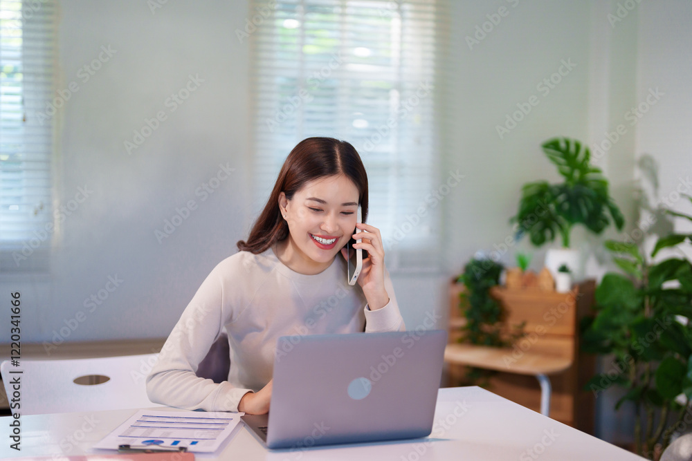 Obraz premium A woman is multitasking, talking on the phone while using her laptop and reviewing documents on the desk.