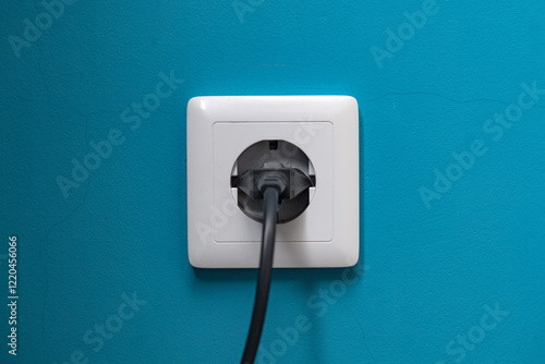 Black power plug is plugged into a white socket on a cyan-painted wall.