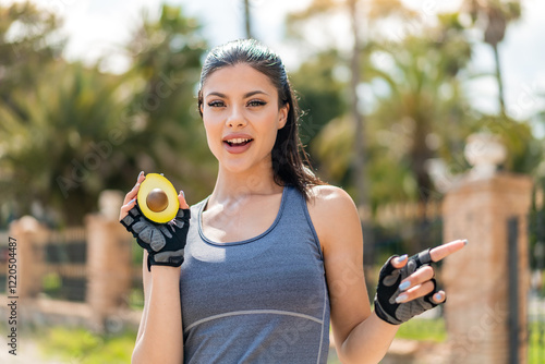 Young pretty sport woman holding an avocado at outdoors surprised and pointing side photo