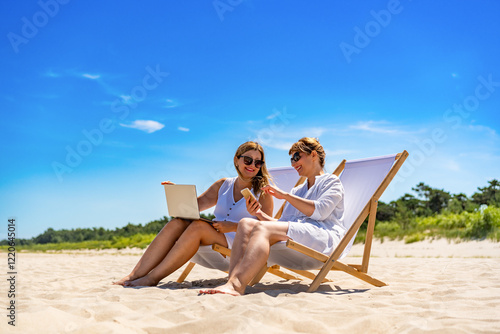 Remote work on beach. Two middle aged women sitting on deckchairs using laptop and smartphone on sandy beach on sunny day