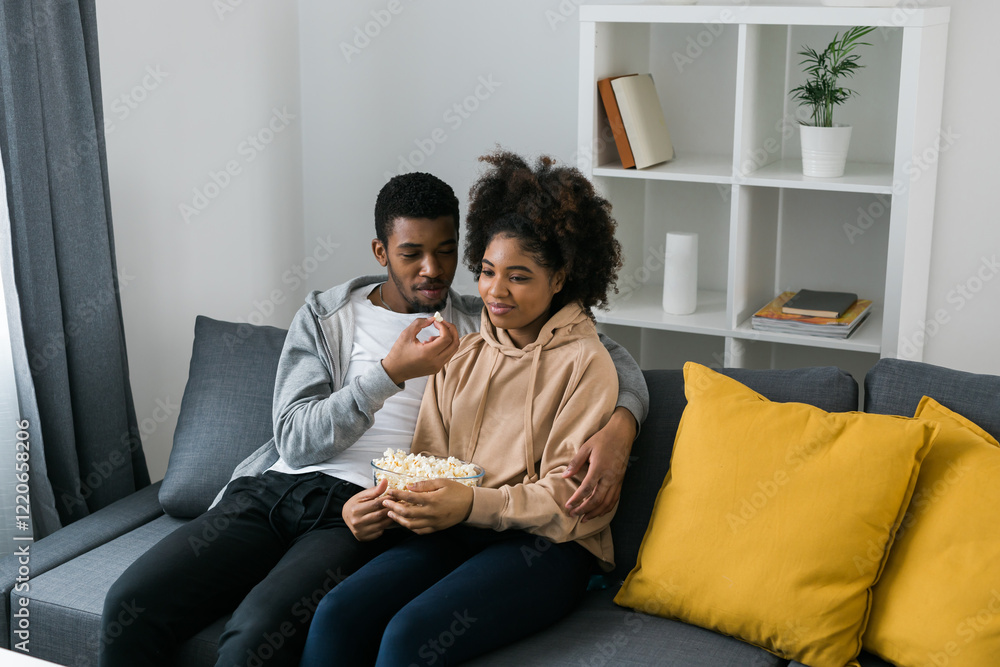 African American couple shares a playful and relaxed moment on the couch, with the man feeding popcorn to the woman. TV, series and streaming service concept African American couple shares a playful and relaxed moment on the couch, with the man feeding popcorn to the woman. TV, series and streaming service concept
