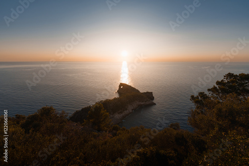 A wide-angle shot of Sa Foradada rock formation bathed in golden sunlight, surrounded by lush greenery and the endless Mediterranean Sea. A must-visit spot in Mallorca.