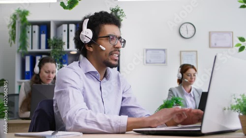 Wallpaper Mural young black man in glasses with headset talking on video call with client and providing assistance online sitting in office with colleagues in background Torontodigital.ca