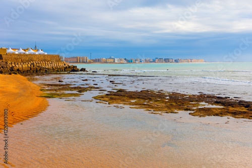 Coastal view of Cadiz city with the Atlantic Ocean in the foreground. Rocky shoreline with shallow water, while the cityscape and a cloudy sky are visible in the distance