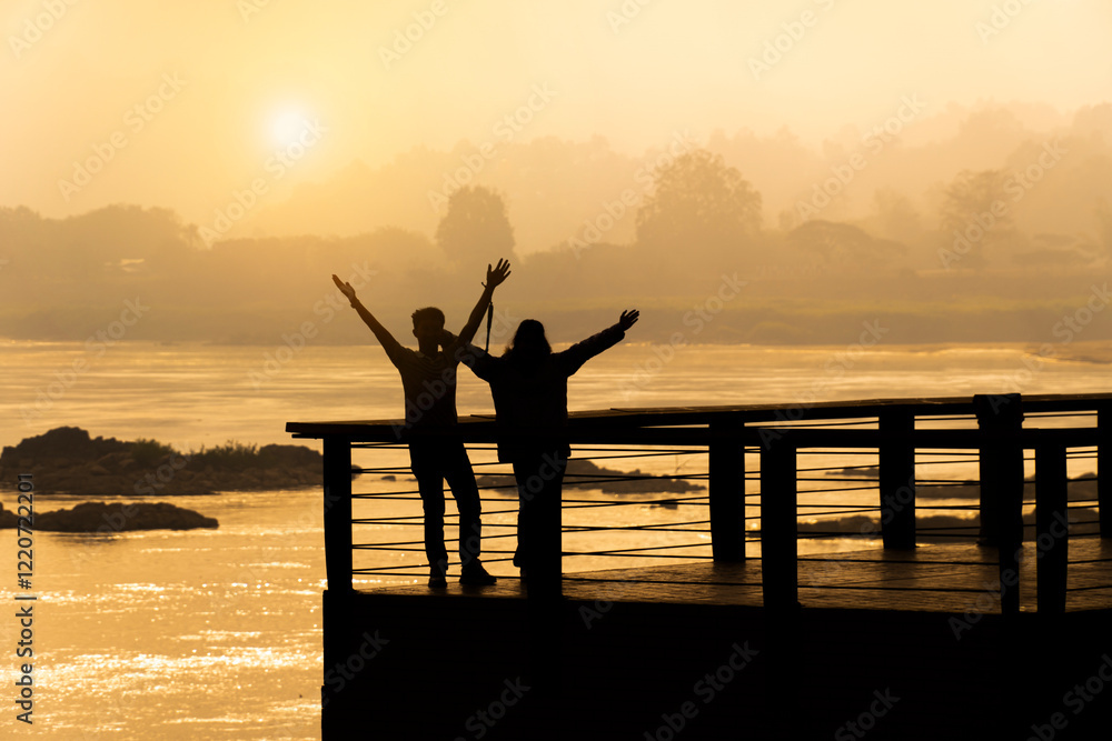 Fototapeta premium Silhouette of a happy couple raising arms on a riverside deck during golden sunset, romantic outdoor scene