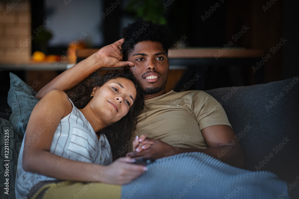 Fototapeta premium A happy couple relaxing on the couch at home, cuddling while watching TV. The woman rests her head on her partner's shoulder as they enjoy a cozy and intimate moment together