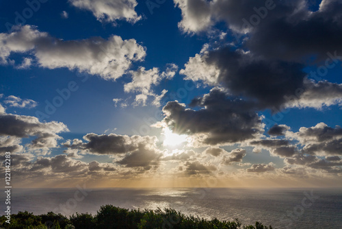 The sun breaks through dramatic clouds over a calm ocean, casting a golden light on the water.