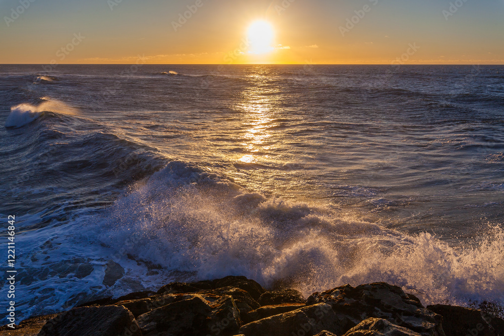 Obraz premium Waves crash against a rocky shoreline at sunset, with the sun setting over the ocean horizon.