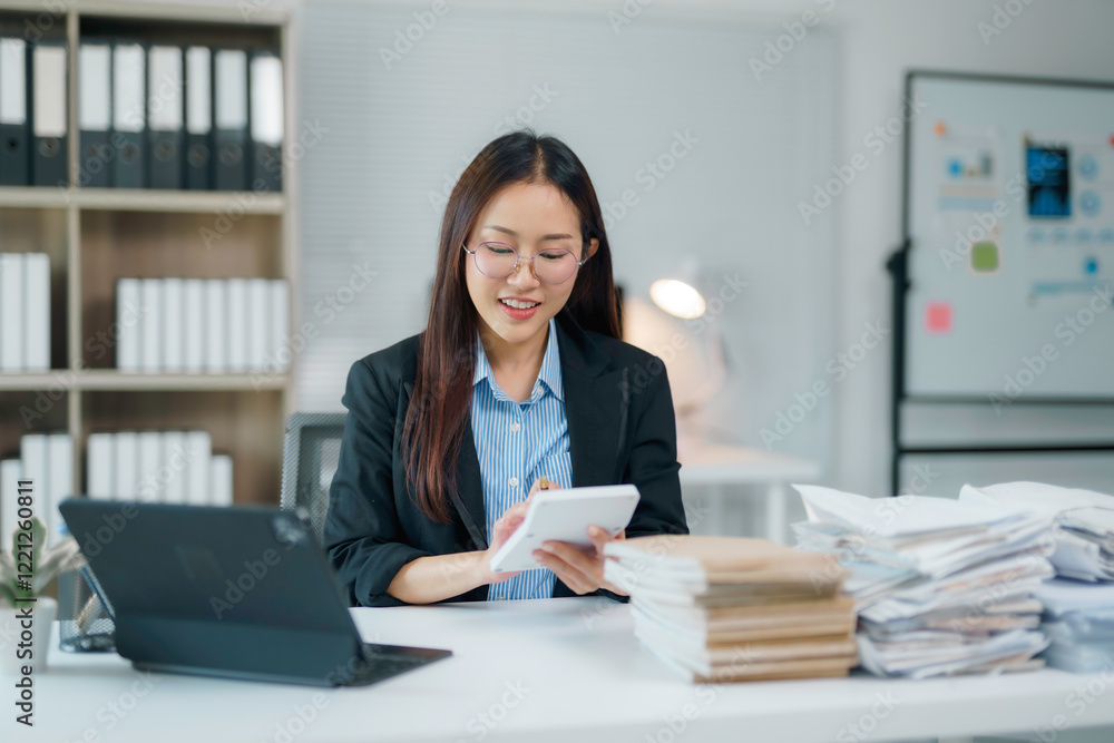 Asian businesswoman calculating and taking notes in office with stacks of paperwork and laptop Asian businesswoman calculating and taking notes in office with stacks of paperwork and laptop