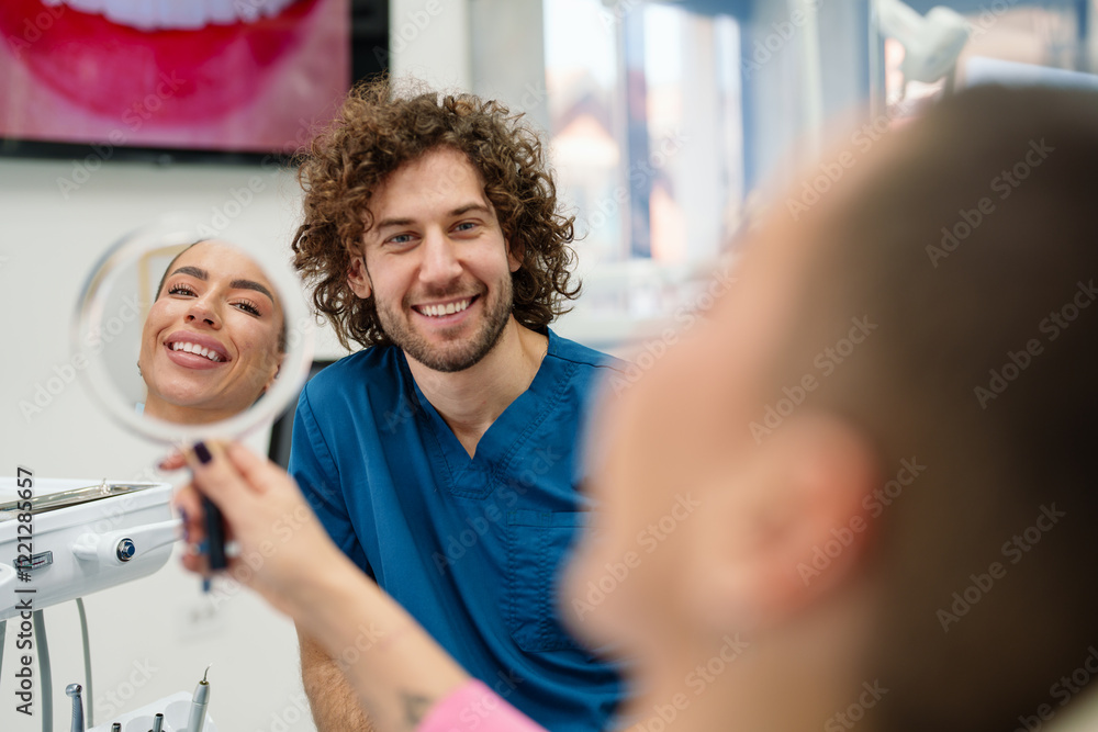 A Caucasian woman smiles while viewing her reflection in a mirror at a dental office. A Caucasian male dentist in blue scrubs stands nearby, indicating a successful dental procedure. A Caucasian woman smiles while viewing her reflection in a mirror at a dental office. A Caucasian male dentist in blue scrubs stands nearby, indicating a successful dental procedure.