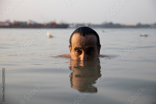 An Indian man taking a holy dip in the river ganges during early morning. he is chest deep submerged in water and is offering prayers to god.