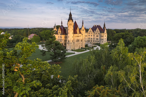 Majestic Moszna Castle on a warm summer morning, surrounded by lush greenery and glowing in soft golden sunlight, reflecting a fairytale-like ambiance perfect for travel and architecture themes.