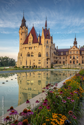 Majestic Moszna Castle on a warm summer morning, surrounded by lush greenery and glowing in soft golden sunlight, reflecting a fairytale-like ambiance perfect for travel and architecture themes.
