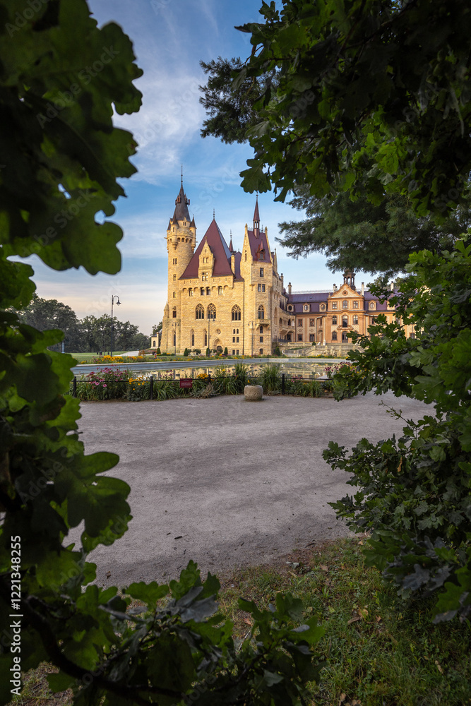 Majestic Moszna Castle on a warm summer morning, surrounded by lush greenery and glowing in soft golden sunlight, reflecting a fairytale-like ambiance perfect for travel and architecture themes.