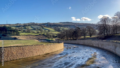 A tranquil rural scene with Swinsty Reservoir spillway framed by trees and sunny blue skies - Nr Harrogate, North Yorkshire, UK
