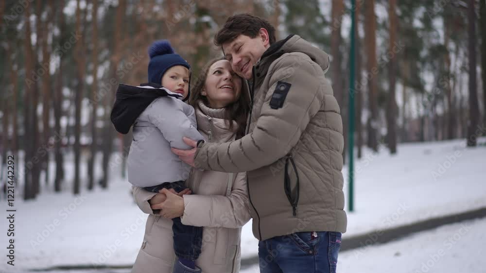 A happy family of three stands together in a snowy park. The father holds the child while smiling warmly at the camera, and the mother stands beside them, also smiling. The winter clothing and snowy A happy family of three stands together in a snowy park. The father holds the child while smiling warmly at the camera, and the mother stands beside them, also smiling. The winter clothing and snowy