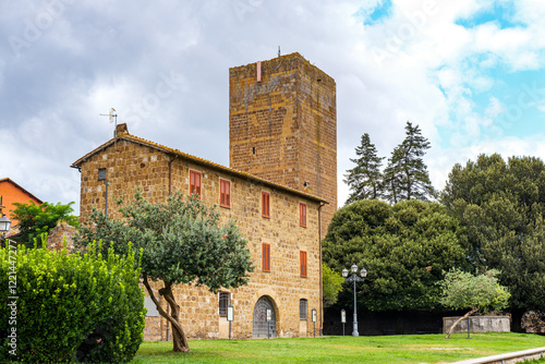 Torre di Lavello, a prominent medieval tower, rises above a historic building in Tuscania, Italy, surrounded by lush vegetation and showcasing the town's architectural heritage under a cloudy sky