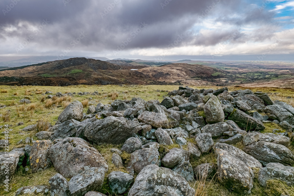 A vast landscape features scattered large rocks on a grassy field, leading to rolling hills in the distance, all set under a cloudy sky during daylight hours. A vast landscape features scattered large rocks on a grassy field, leading to rolling hills in the distance, all set under a cloudy sky during daylight hours.
