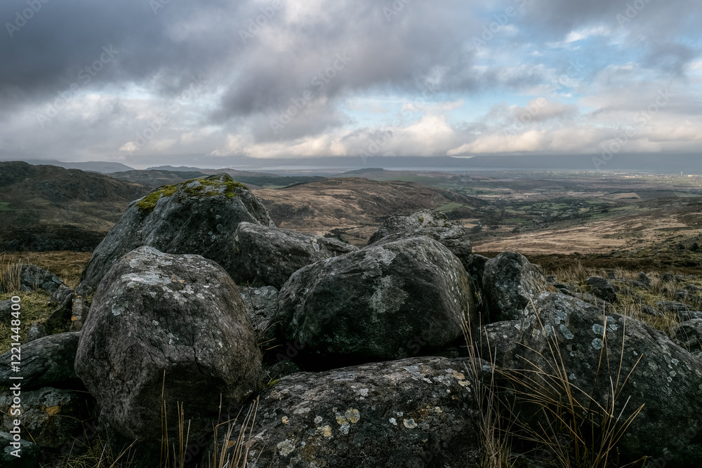 A cluster of large, dark grey rocks, lichen-covered, sits atop a grassy hill. A distant valley and cloudy sky are visible. A cluster of large, dark grey rocks, lichen-covered, sits atop a grassy hill. A distant valley and cloudy sky are visible.