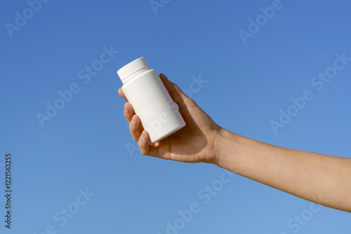 Female hand holding blank mockup pill bottle on blue sky background. Concept of pharmacy, medicine, healthcare and natural vitamins D3, magnesium, calcium, omega