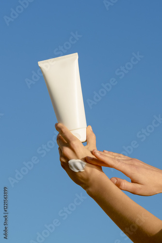 Vertical image of a female hand applying a sample of cream and holding a white mockup tube of moisturizer on a blue sky background. Concept of facial and body skin care, beauty products, aesthetics