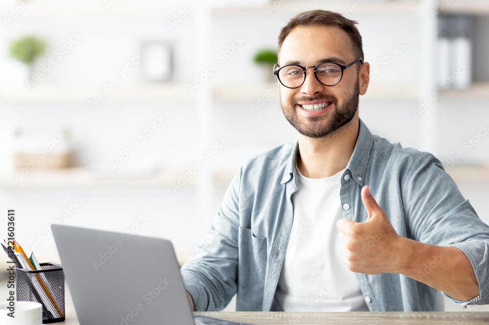 Obraz premium Happy young guy sitting at desk with flag of Turkey, using laptop, showing thumb up gesture at home. Online foreign education, emigration and citizenship, working abroad concept
