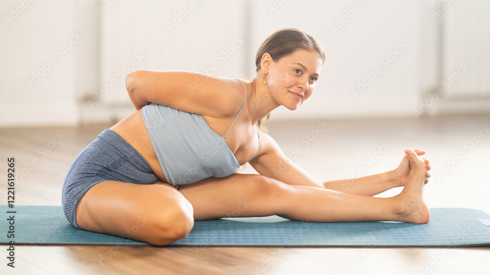 Fototapeta premium Plastic girl training head to knee position on blue mat in yoga studio with wide windows