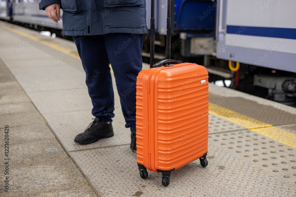 A young man with a small orange suitcase waits for a train at the train station for traveling. Lifestyle, people, adventure and tourism concept.