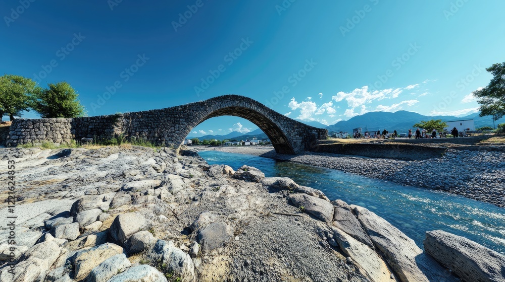 Fototapeta premium A stone bridge arches over a river with mountains in the background under a clear blue sky.
