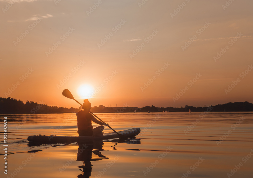 Sunlit silhouette of a women paddling sitting on a standup paddleboarding (SUP) desk. Scenic sunset landscape view at Kiekrz lake (Jezioro Kierskie) near Poznań, Wielkopolskie, Poland.