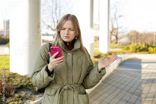 Perplexed woman holding cellphone in hands on the street, looking at screen with irritated expression, made a mistake or annoyed by texts and calls. Negative emotion. High quality photo