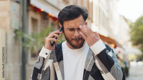 Angry mad stressed Indian young man in casuals arguing while talking on smartphone irritated shout quarrel outdoors. Hispanic offended dissatisfied guy walking on urban city street. Town lifestyles.