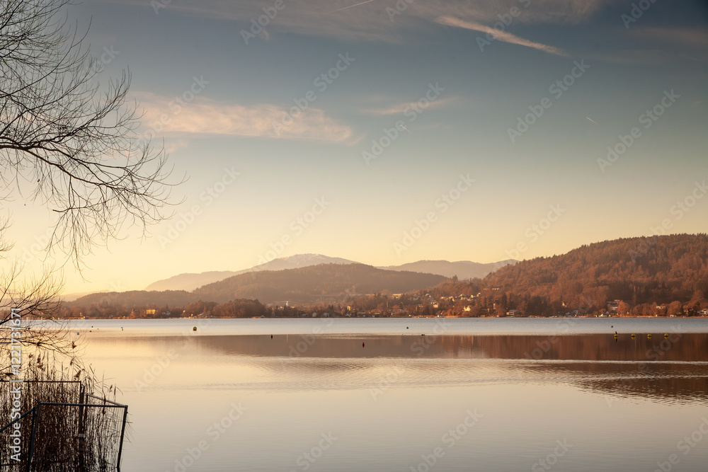 A serene evening view across the Wörthersee reveals calm waters and soft, golden hues, with distant hills framing the tranquil lakeside panorama at dusk. A serene evening view across the Wörthersee reveals calm waters and soft, golden hues, with distant hills framing the tranquil lakeside panorama at dusk.