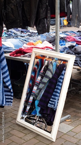 White framed mirror reflects hanging woven clothing at flea market, table with clothing and hanging leather jackets surround mirror. Historic Waterlooplein Market or Markt, Amsterdam, the Netherlands. photo