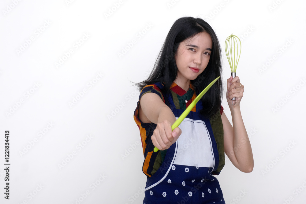 A young woman wearing an apron holds a spatula and a whisk; in a serious expression
