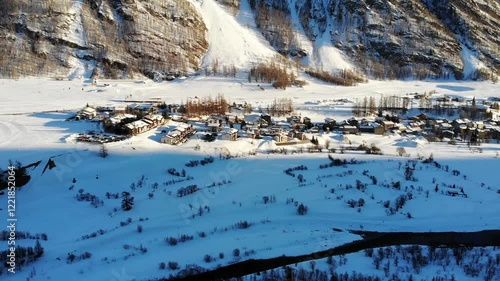 A drone pans upward, revealing a quaint village nestled at the foot of a towering mountain. The snow-covered landscape creates a serene winter scene, showcasing the harmony of the nature