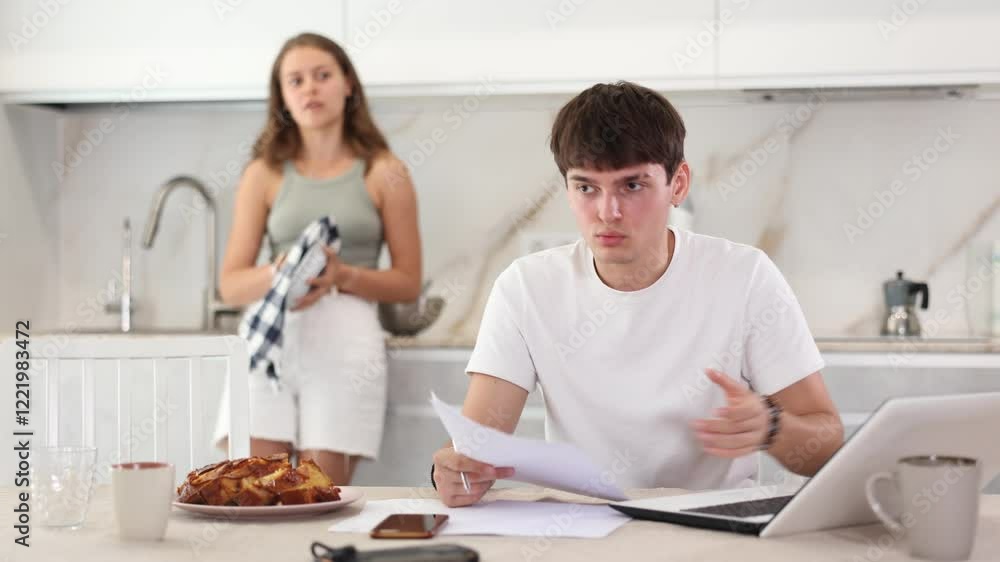 Sad young guy working on laptop while her girlfriend is quarreling with her standing behind in the kitchen Sad young guy working on laptop while her girlfriend is quarreling with her standing behind in the kitchen