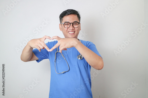 Smiling professional young Asian male doctor or nurse wearing a blue uniform and stethoscope showing a Korean heart gesture isolated on yellow background. Healthcare medicine concept