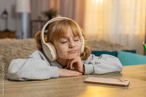 Young girl lying on table listening to an audiobook with headphones, reacting with excitement, surprise. Caucasian child smiles joyfully, wondering what happens next in story, enjoys adventure at home photo