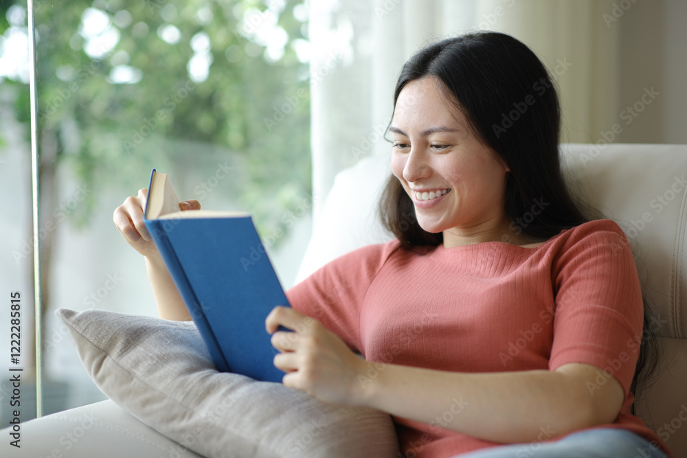Obraz premium Happy asian woman reading a paper book on a couch
