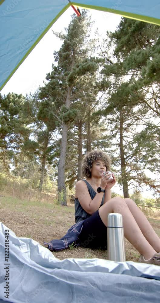 Vertical video: Camping in forest, woman enjoying hot drink outside tent with thermos nearby Vertical video: Camping in forest, woman enjoying hot drink outside tent with thermos nearby