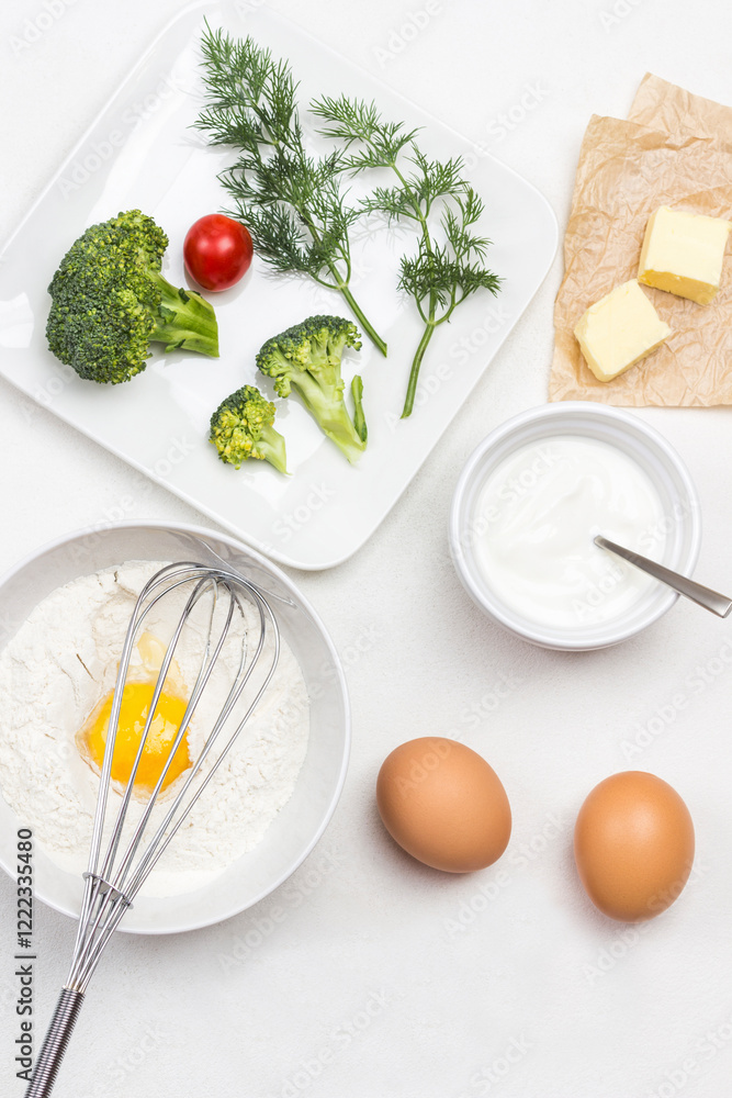 Broken egg with flour in bowl. Whisk on bowl. Butter on paper. Broccoli, tomatoes and dill