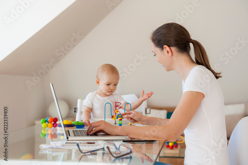 Mother using laptop and little daughter playing at table at home photo