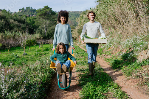 Family walking on a dirt track, pushing wheelbarrow, carrying crate with vegetables photo