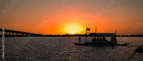 Silhouette of a traditional Philippine boat filled with passengers, floating on calm waters during a vibrant sunset, creating a serene and picturesque scene