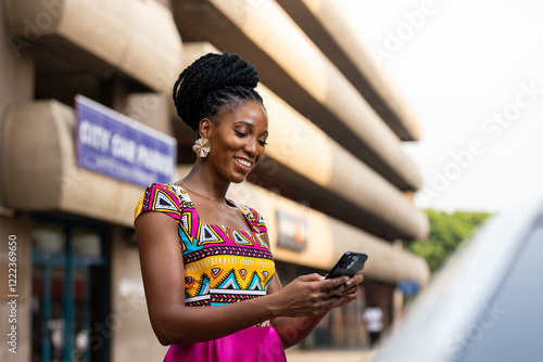 Smiling African woman in vibrant African print dress checks her phone outdoors.