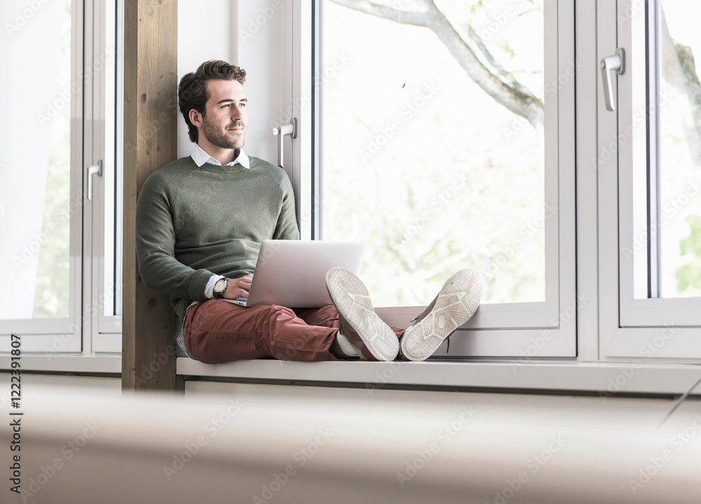 Young businessman sitting on windowsill, using laptop Young businessman sitting on windowsill, using laptop